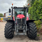 Front view of a red New Holland tractor with large rear tires in a parking lot, hedge on the right and a building in the background.