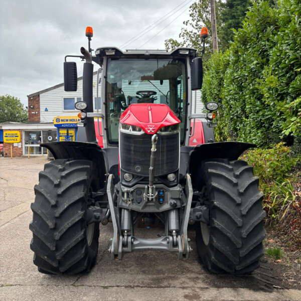 Front view of a red New Holland tractor with large rear tires in a parking lot, hedge on the right and a building in the background.