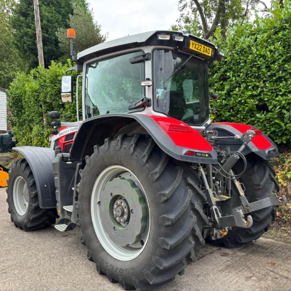 Red Massey Ferguson tractor with oversized rear tires parked in a yard, hedge and building in the background, orange beacon on the roof, and a yellow attachment on the ground to the left.] ,