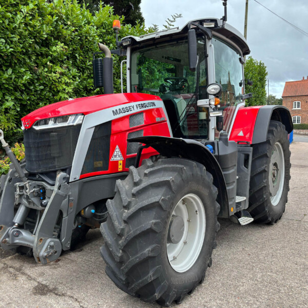 Red Massey Ferguson tractor parked on a street beside a green hedge and a brick building in the background.