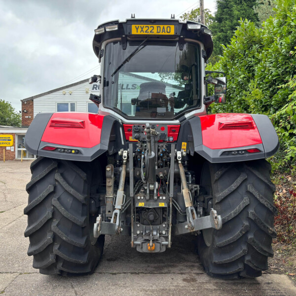Front view of a large red tractor with oversized rear tires, parked in a dealership lot with a hedge on the right and a building in the background.