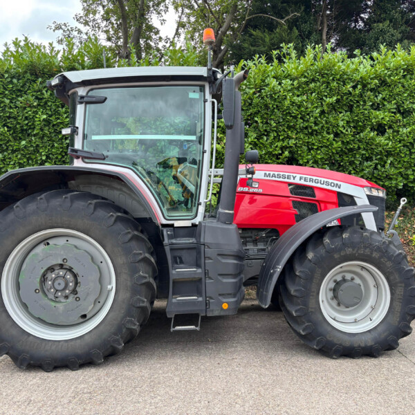 Red Massey Ferguson tractor with oversized tires parked in front of a tall green hedge by a paved surface.