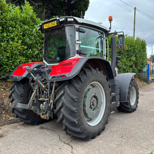Front-left view of a red and gray tractor parked beside a tall green hedge on a suburban street, with large tires visible.