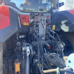 Close-up of a heavy machinery cab control panel with many levers, hoses, and hydraulic lines, red bodywork visible on the sides.