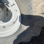 Close-up of a large off-road tire and wheel with dirt and rusted lug nuts on rocky ground.