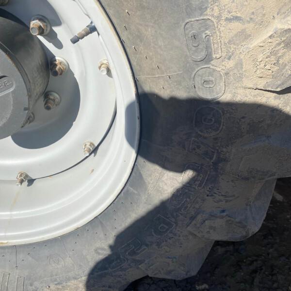 Close-up of a large off-road tire and wheel with dirt and rusted lug nuts on rocky ground.