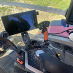 Close-up of a tractor cab control console with a mounted touchscreen tablet, a multi-button joystick, and numerous controls, with a gravel and grass backdrop outside.