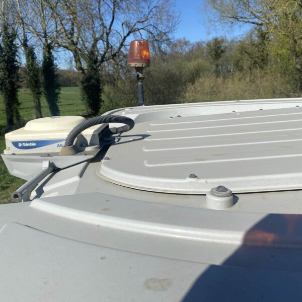 Close-up of an amber warning beacon mounted on a farm vehicle roof, with green fields and bare trees in the background.