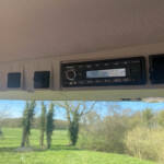 Vehicle dashboard radio unit visible at top of image with a landscape of green field and trees through the windshield, seen inside a car or van