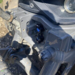 Close-up of a heavy machinery front panel showing a hydraulic hose, exposed bolt, and dusty surface with mud on tires nearby.