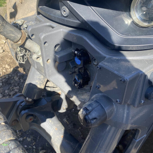 Close-up of a heavy machinery front panel showing a hydraulic hose, exposed bolt, and dusty surface with mud on tires nearby.