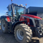 Red Massey Ferguson tractor parked on a dirt lot beside a green metal building; large tires and equipment attached.