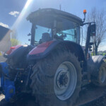 Front of a large tractor with huge rear wheel, orange beacon on top, parked outdoors on a sunny day.