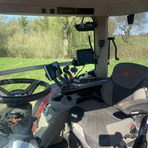 Interior view of a tractor cab with steering wheel, multiple screens, and control panels; green fields and trees outside through the windows at a rural site.