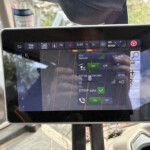 Touchscreen tablet in a vehicle showing a detailed navigation/map screen and status icons on the display. Background shows a bottle and cab interior.