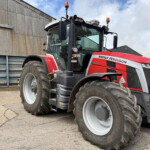 Red Massey Ferguson tractor with large front tires parked beside a corrugated metal building, front view showing cabin and orange beacon lights.
