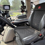 Interior of a truck cab showing a steering wheel, multiple screens, and mounted devices near the dashboard; patterned driver seat with a red logo.