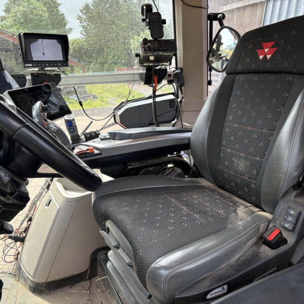 Interior of a truck cab showing a steering wheel, multiple screens, and mounted devices near the dashboard; patterned driver seat with a red logo.