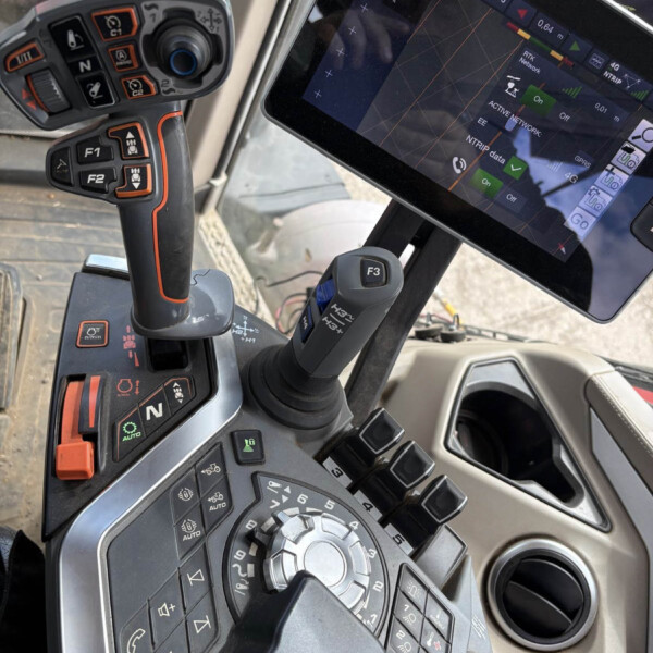 Close-up of a heavy machinery cockpit with a joysticky controller, numerous illuminated buttons, and a mounted touchscreen display showing control data on the right side.