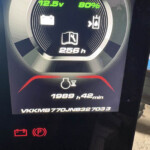 Circular car dashboard gauge showing 12.5V and 80% with a book icon and 256 h; warning icons below indicate battery and parking brake, durée 1989 h 42 min