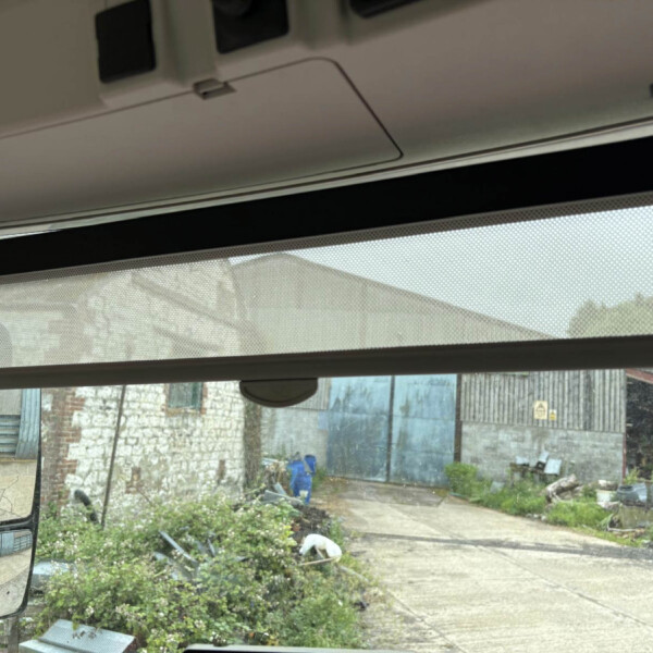 Interior of a vehicle looking out through a dusty windshield at a yard with a brick wall and metal fencing; side mirror visible on the left.