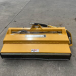 Yellow industrial hydraulic attachment on a concrete surface, showing hoses and mounting frame with warning labels visible.