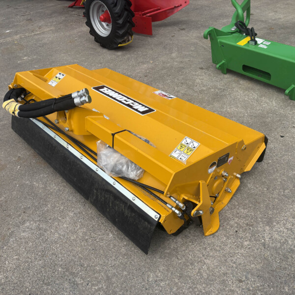 Yellow industrial mower attachment with hydraulic hoses lying on asphalt, nearby green farm equipment visible as a backdrop.