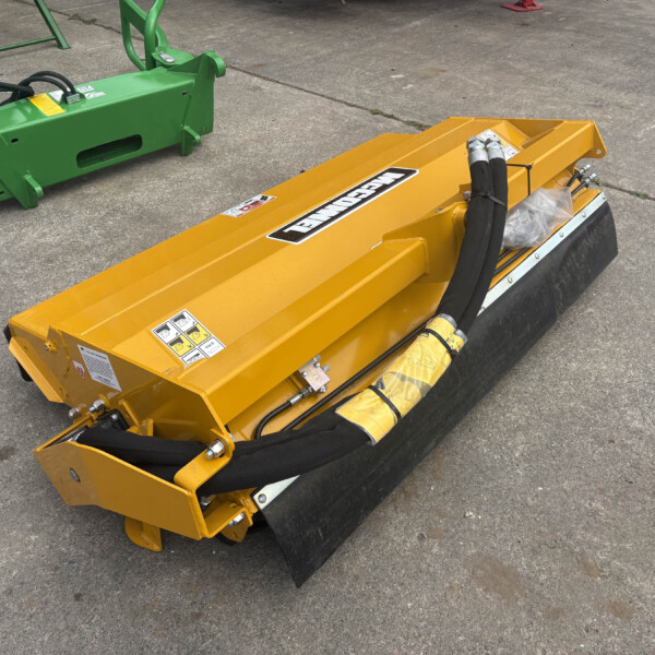 Yellow trenching attachment with hydraulic hoses on a concrete surface, nearby there is green construction equipment.