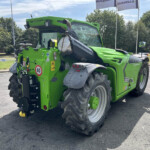 Green agricultural harvester viewed from the side, showing large rear tires, warning labels, and a long cutting header; sunny day with trees in background.
