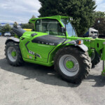 Bright green telehandler with a large front lifting arm, parked in a paved lot beside a building and trees, with a white truck in the background.