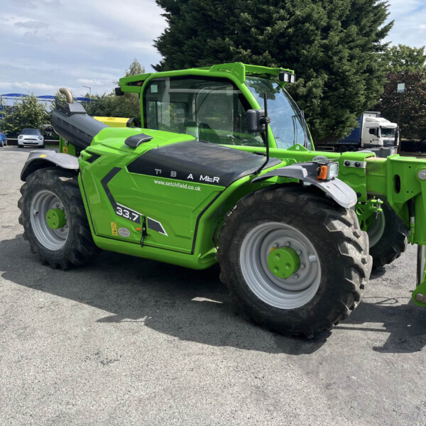 Bright green telehandler with a large front lifting arm, parked in a paved lot beside a building and trees, with a white truck in the background.