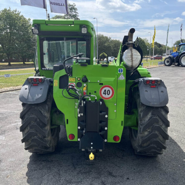 Rear view of a large green tractor with oversized tires and hitch, parked on a paved lot with blue tractors in the background.