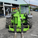 Bright green agricultural tractor with frontloader forks facing the camera, parked outside a dealership showroom with glass windows and other vehicles in the lot