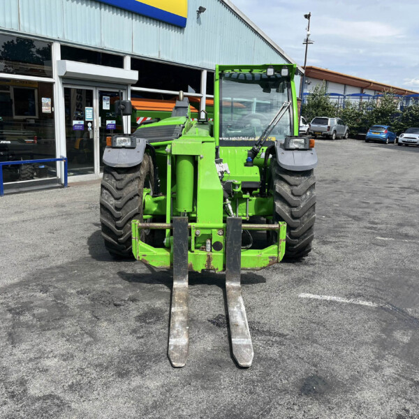 Front view of a bright green agricultural tractor with fork attachments, parked outside a service shop with glass doors and windows behind it.