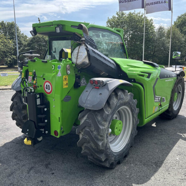 Green agricultural harvester viewed from the side, showing large rear tires, warning labels, and a long cutting header; sunny day with trees in background.