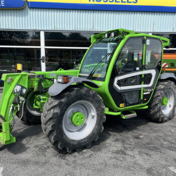 Bright green utility tractor with a front loader attachment parked outside a building with large glass windows and a blue-yellow sign above it, indicating a dealership exterior.