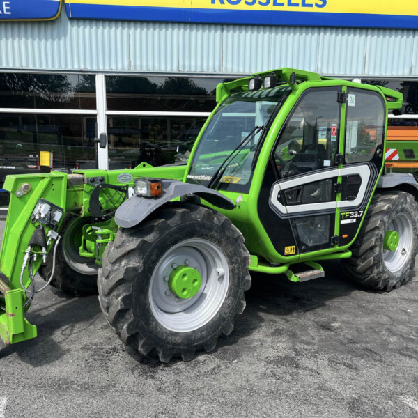 Bright green utility tractor with a front loader attachment parked outside a building with large glass windows and a blue-yellow sign above it, indicating a dealership exterior.