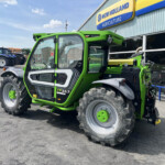 Bright green New Holland tractor in foreground outside a dealership, with blue tractors lined up in the background and a 'New Holland Agriculture' sign above.
