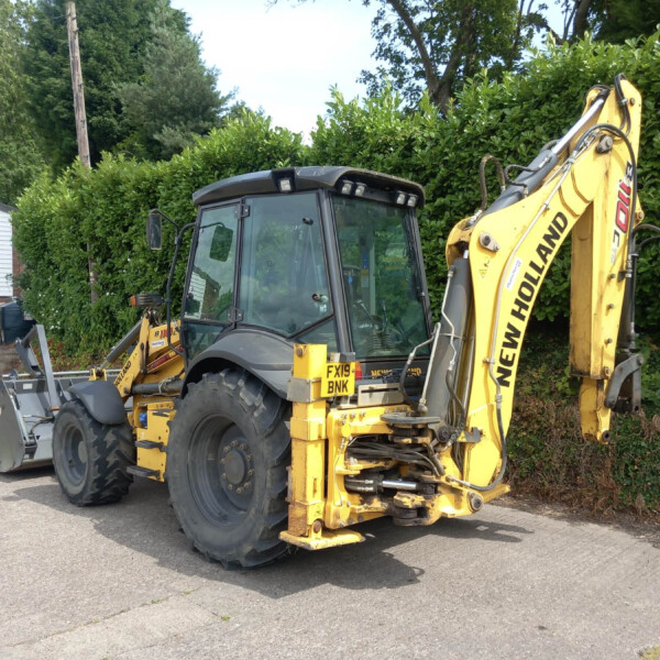 Yellow backhoe loader parked on a paved lot beside a tall green hedge and a white building. construction vehicle awaiting use in a yard.