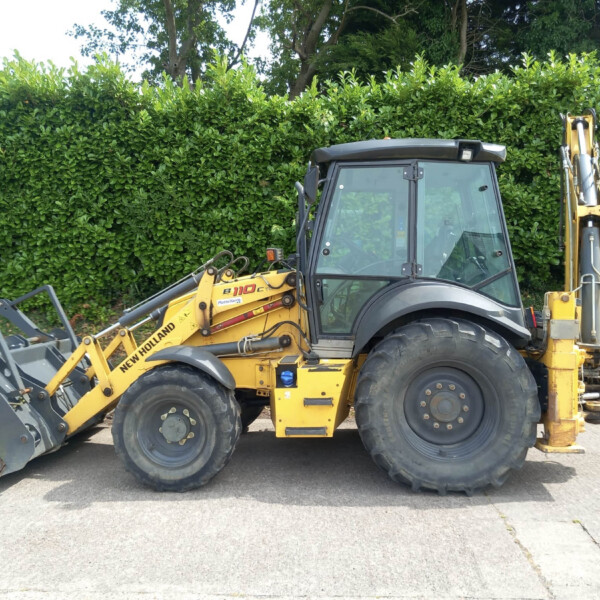 Backhoe loader parked on a paved surface in front of a tall green hedge, showing a front bucket and hydraulic machinery (New Holland B110C).