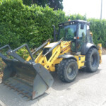 Yellow backhoe loader with a front bucket parked along a road beside a tall green hedge.