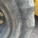 Close-up of a large tire's dusty sidewall with embossed numbers along the edge and a yellow vehicle frame in the background.