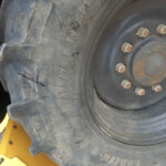 Close-up of a large off-road tire on a metal wheel with rusted lug nuts, showing dirt and cracked tread near the yellow machinery frame in the foreground.