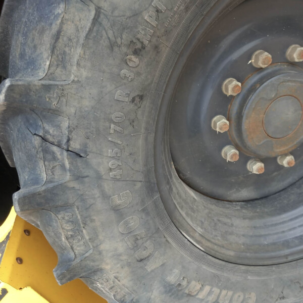 Close-up of a large off-road tire on a metal wheel with rusted lug nuts, showing dirt and cracked tread near the yellow machinery frame in the foreground.