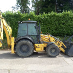 Yellow backhoe loader parked on a paved surface with its bucket bucketed down, in front of a green hedge.