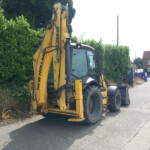 Yellow backhoe loader parked beside a hedge on a street, with a brick building in the background.