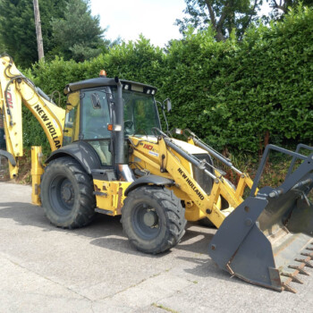Yellow New Holland backhoe loader parked on a paved surface beside a dense green hedge.