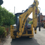 Yellow New Holland excavator on a street beside a hedge, with visible hoses and a rear license plate FX19 BNK.