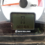Close-up of a New Holland tractor dashboard: red needle gauge, digital display with an hourglass icon and numeric readout.
