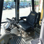 Inside tractor cab showing steering wheel and driver's seat, with multiple blue tractors parked outside in a lot